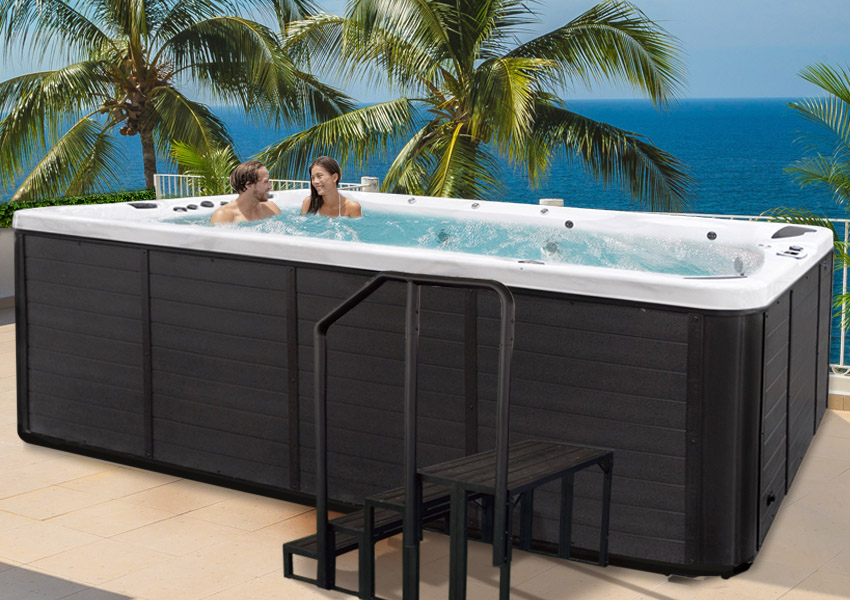 Two people laughing in a Cal Spas Swim Pro™ hot tub, with steps, on a patio with palm trees and ocean in the background.